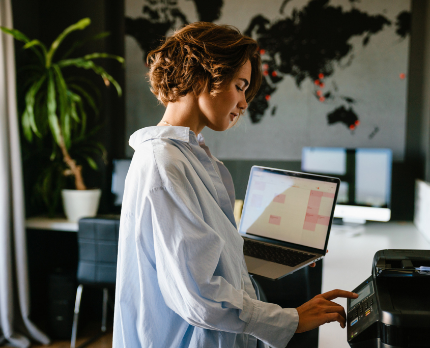 Businesswoman Holding Laptop And Pressing Button On Panel Of Printer In Office