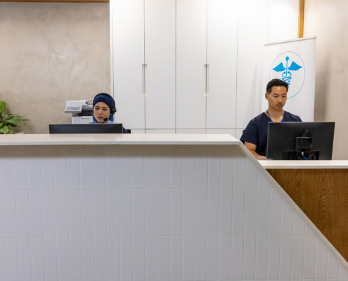 Healthcare Staff Speaking Into Headset At Clinic Desk With Monitors